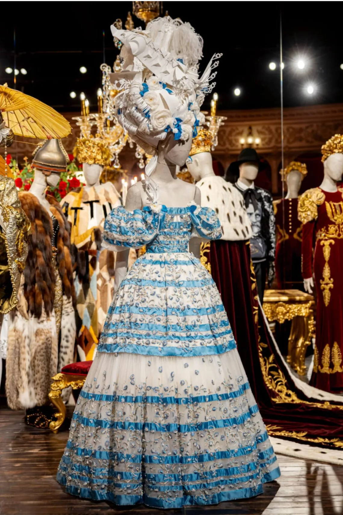 Mavi-beyaz tonlarda, kabarık etekli ve heykelsi baş aksesuarıyla Dolce & Gabbana Alta Moda elbisesi; tarihsel siluetler ve el işçiliğinin buluştuğu sergi sahnesi.