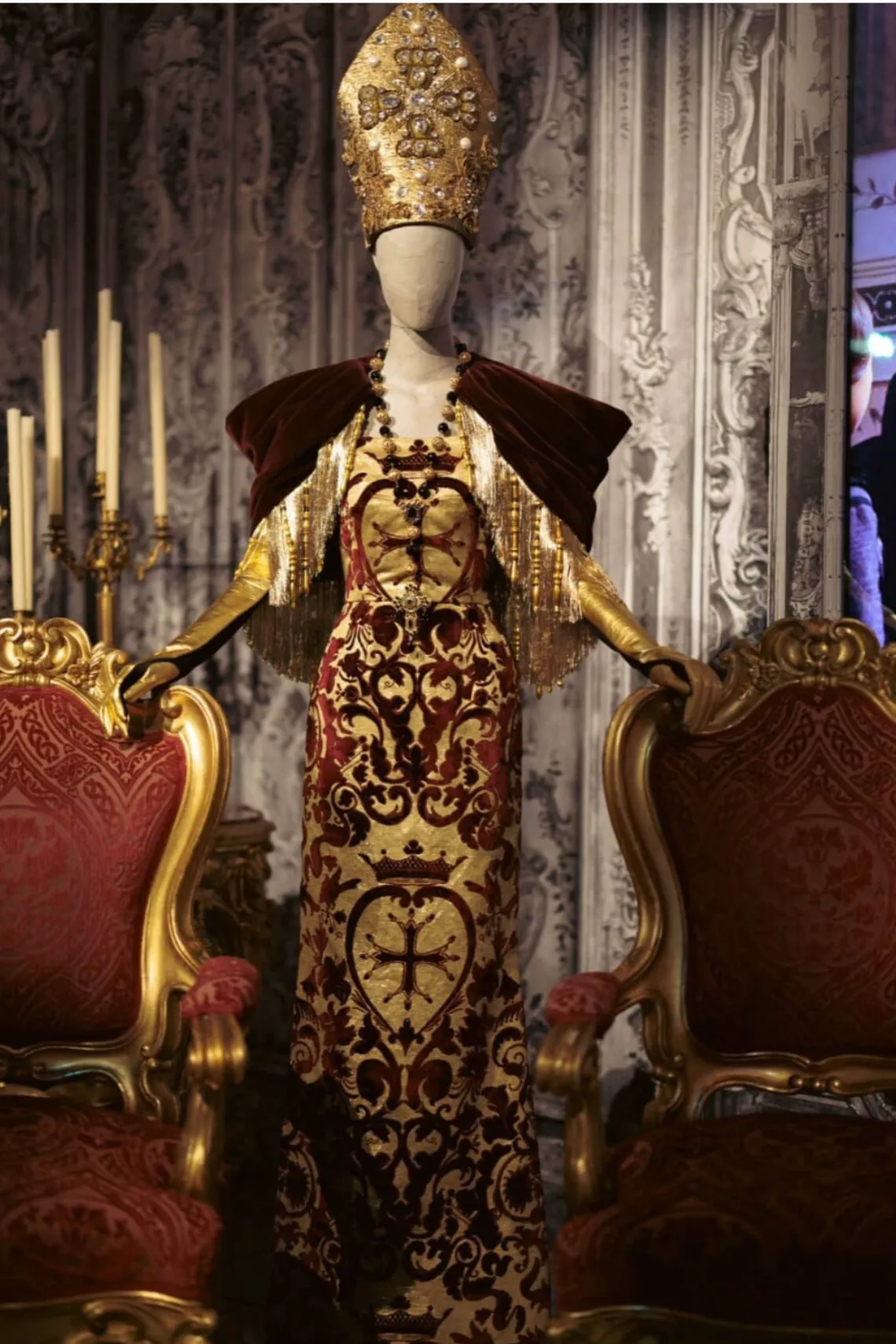 Altın ve bordo renk paletiyle tasarlanan, dini motifler ve işlemelerle bezeli Dolce & Gabbana Alta Moda elbisesi; barok iç mekânla bütünleşen dramatik kompozisyon.