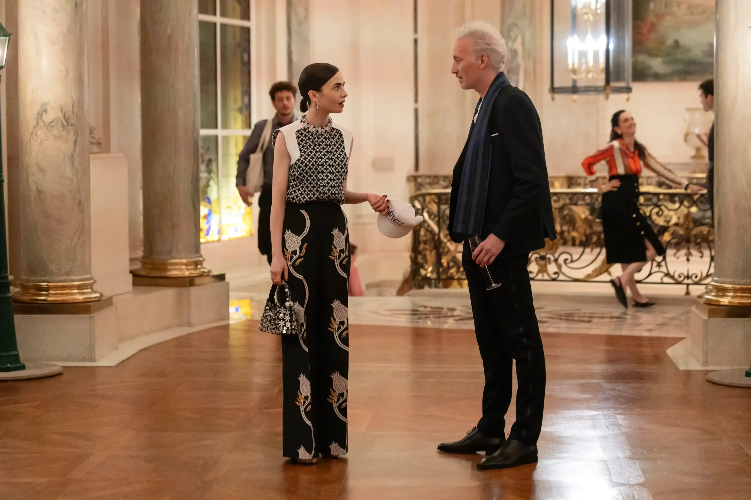 Emily in Paris dizisinde tarihi bir salonda düzenlenen davette siyah desenli elbise ve klasik erkek takımıyla zarif karşılaşma sahnesi.
