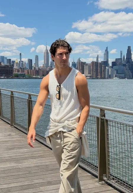 New York silüeti önünde, bej pantolon ve beline bağladığı açık renk bir kıyafetle tamamladığı beyaz V yaka kolsuz tişörtüyle yürüyen genç bir erkek, günümüz erkek moda markalarının rahat ve şık tarzını yansıtan modern bir görünüm sergiliyor.