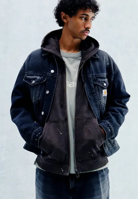 Koyu renk kıvırcık saçlı ve bıyıklı genç bir adam, sağa bakarken, Carhartt WIP markasının yıpranmış detaylı koyu denim ceketini, üzerine beyaz boya lekeleri olan kapüşonlu gri bir sweatşörtle ve açık renk bir tişörtle katmanlı bir şekilde giymiş, elleri ceplerinde durarak modern erkek modası için karakteristik bir tarz sergiliyor.