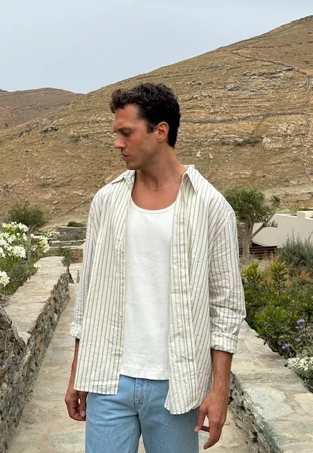Kıvırcık saçlı genç bir adam, açık renk kot pantolonu, beyaz atleti ve üzerine açık bırakılmış çizgili, bol kesim gömleğiyle, taş patikalı ve kuru tepelerin olduğu doğal bir ortamda profilden duruyor; bu rahat ve şık tarz, en iyi erkek moda markalarının ilham veren koleksiyonlarını temsil ediyor.