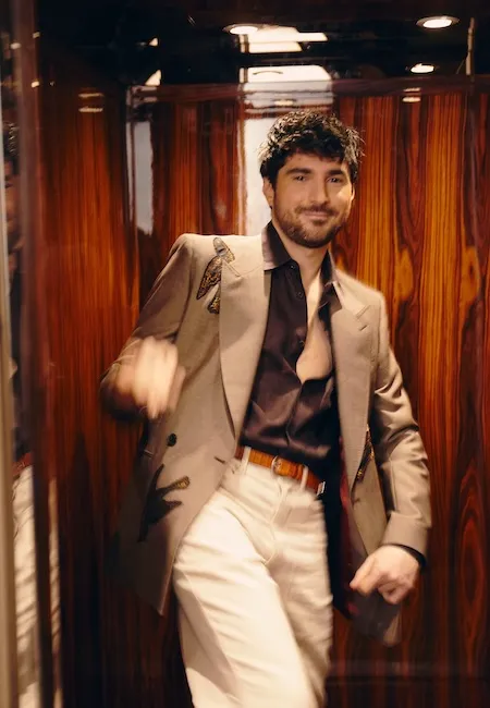 Koyu kıvırcık saçlı ve sakallı, gülümseyen bir adam, üzerinde koyu renk kelebek veya böcek işlemeleri bulunan bej bir blazer ceket, koyu renk açık yakalı bir gömlek, açık renk pantolon ve kahverengi bir kemerle parlak ahşap panelli bir ortamda şık bir şekilde durarak kameraya bakıyor; bu görünüm en iyi erkek moda markalarının özgün tasarım anlayışını vurguluyor.