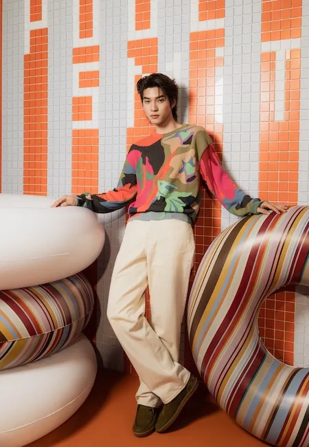 Çok renkli, soyut desenli bir süveter, açık renk pantolon ve koyu yeşil mokasen giyen genç bir erkek model, Paul Smith'in ikonik çizgili desenine sahip şişme halkalar ve turuncu-gri kare fayanslı modern bir arka plan önünde, en iyi erkek moda markalarından Paul Smith tasarımını dikkat çekici bir şekilde sergiliyor.