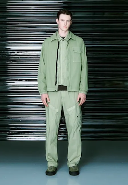 Metalik panelli bir arka plan önünde duran genç erkek model, Stone Island markasının modern estetiğini yansıtan, açık yeşil renkte fermuarlı bir ceket ve uyumlu kargo pantolon kombinasyonunu taşıyarak en iyi erkek moda markalarından birinin stilini öne çıkarıyor.
