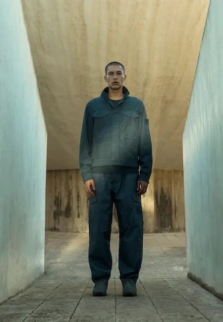 Kısa saçlı genç bir adam, beton dokulu dar bir koridorda dururken, Stone Island markasının sol kolunda logosu olan, koyu petrol mavisi bir ceket ve aynı renk kargo pantolondan oluşan modern ve minimalist bir erkek moda kombinini sergiliyor.