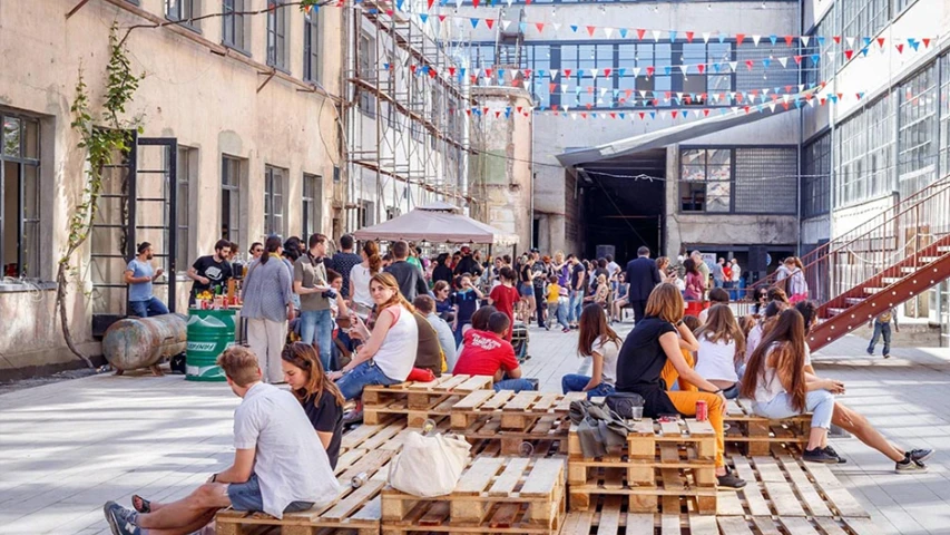 Büyük bir kalabalık, kırmızı, beyaz ve mavi üçgen bayraklarla süslenmiş, eski fabrika binalarının çevrelediği geniş bir avluda, ahşap paletlerden yapılmış oturma alanlarında sohbet edip güneşli havanın tadını çıkararak bir araya gelmiş.