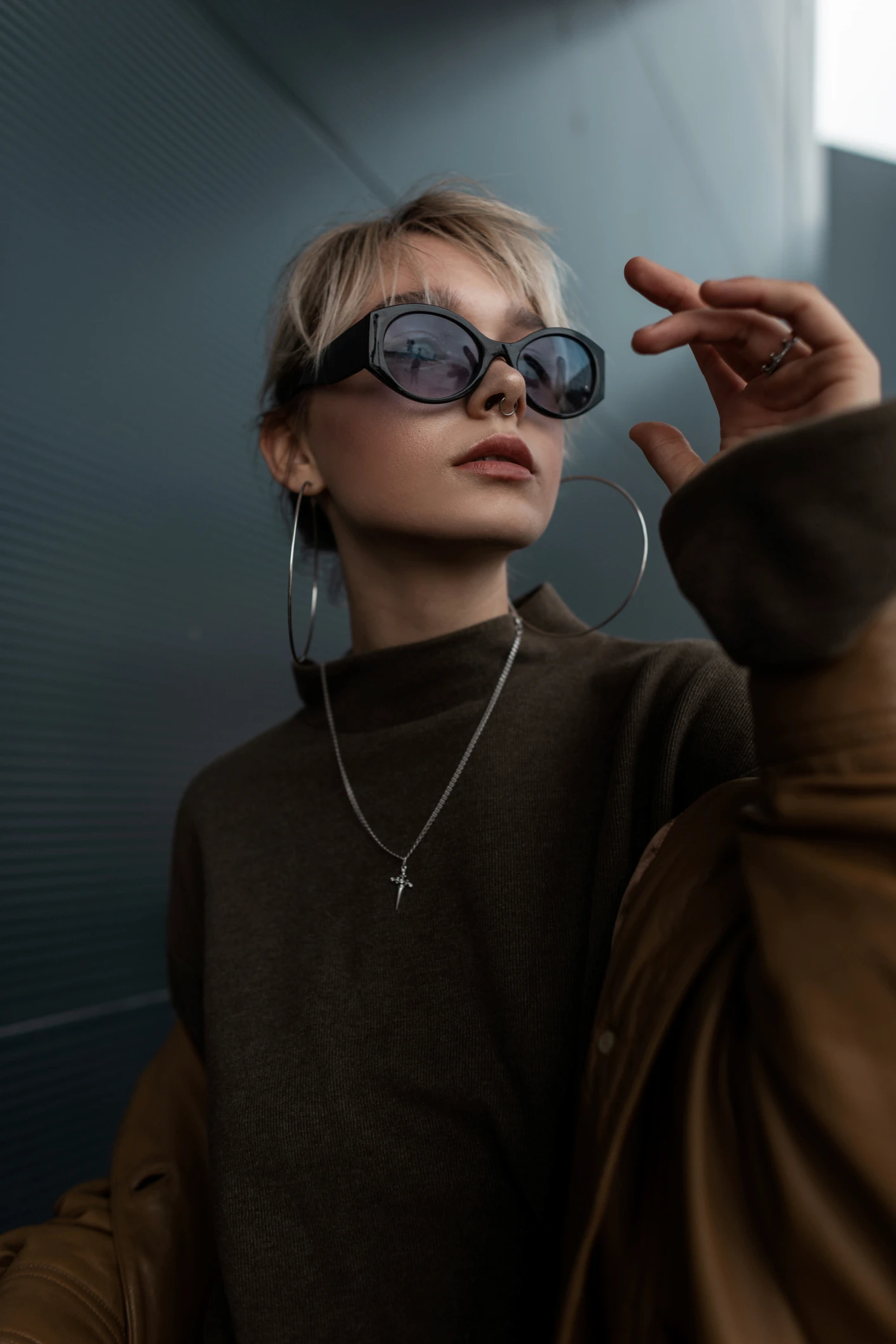 Kısa sarı saçlı ve septum piercingli, cesur, büyük siyah güneş gözlüğü, iri gümüş halka küpeler, koyu yeşil boğazlı kazak ve kahverengi deri ceket giyen şık genç bir kadın, kişisel tarzı yansıtan güneş gözlüğü seçerken göz önünde bulundurulabilecek çağdaş bir estetiği vurgulayarak saçını veya gözlüğünü hafifçe ayarlarken yukarı doğru bakıyor.