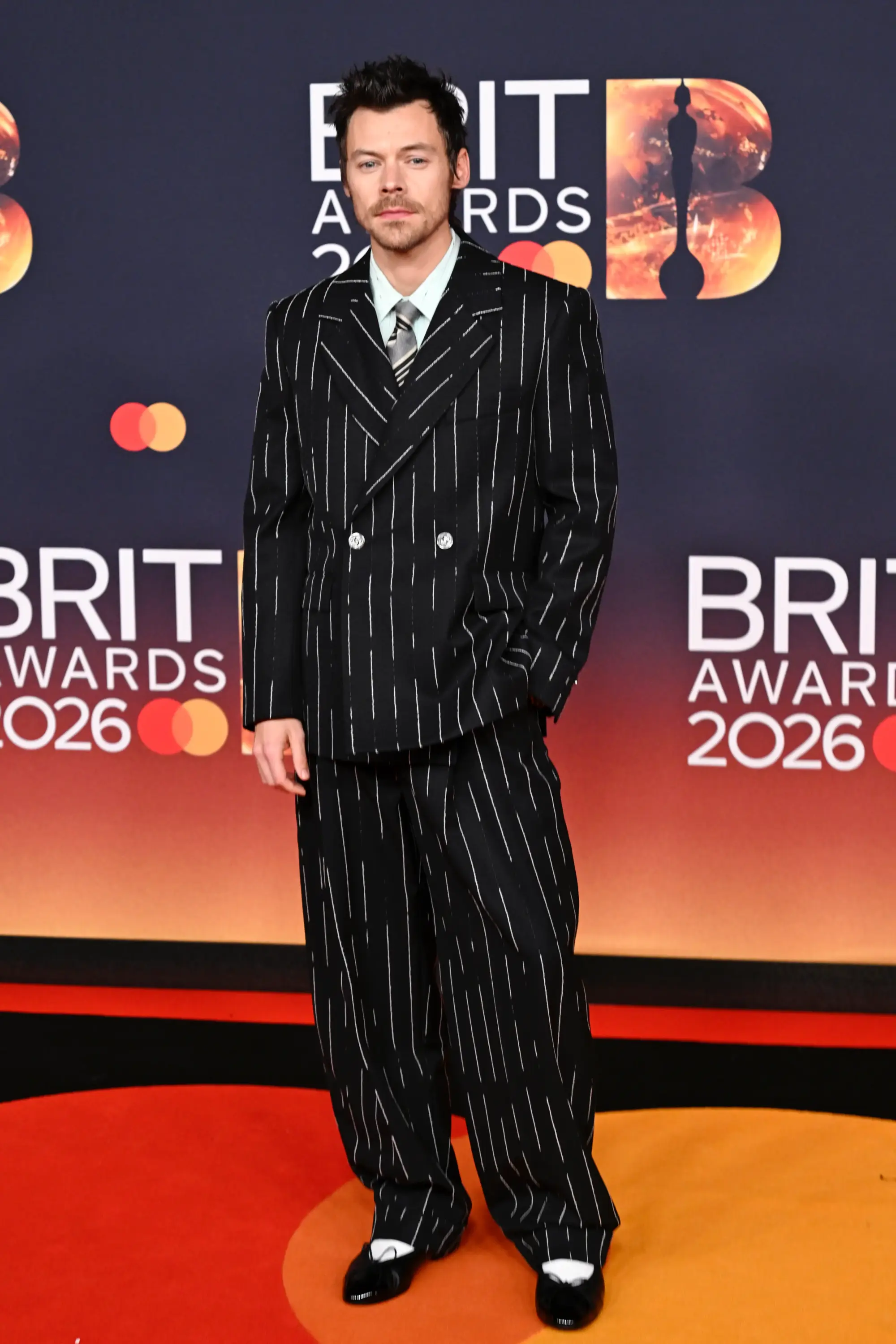 Harry Styles, BRIT Awards 2026 kırmızı halısında ince çizgili siyah çift düğmeli takım elbise ve kravat kombinasyonu ile klasik terziliği modern bir tavırla yorumlarken; geniş paça pantolon ve kontrast ayakkabı detayıyla güçlü ve sofistike bir erkek silueti sergiliyor.