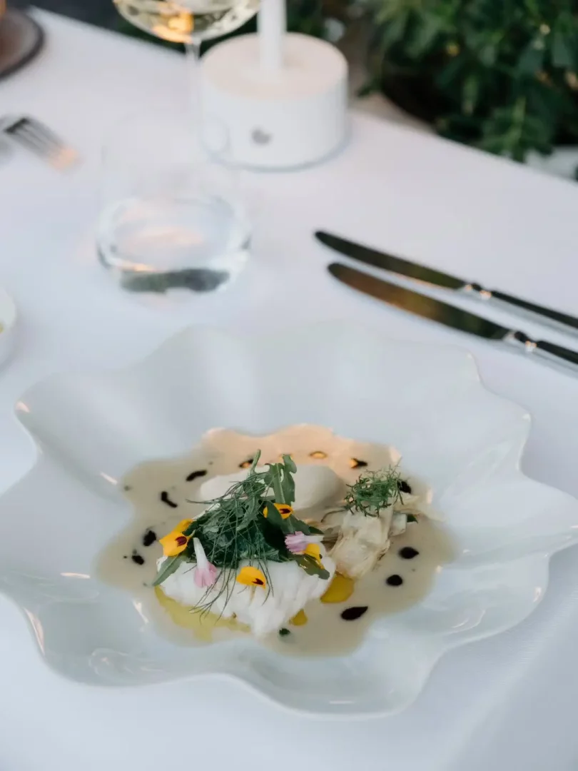 Muğla'nın Michelin Yıldızlı restoranlarından biri olan Kitchen Bodrum'un zarif sunumuyla dikkat çeken bir tabağında, beyaz etli balık mevsimsel yeşillikler ve yenilebilir çiçeklerle süslenmiş, modern soslarla çevrili olarak beyaz bir masa üzerinde görülüyor, bu da Ege mutfağının yaratıcı dokunuşlarını yansıtıyor.