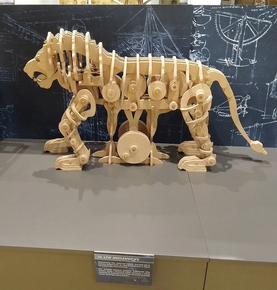 Leonardo da Vinci'nin dahiyane icatlarından biri olarak sergilenen, hareketli parçaları ve dişlileri görünen, doğal ahşap renginde, yan profilden çekilmiş karmaşık bir mekanik aslan modeli, arkasında mühendislik çizimlerinin olduğu koyu renkli bir fonun önünde duruyor.
