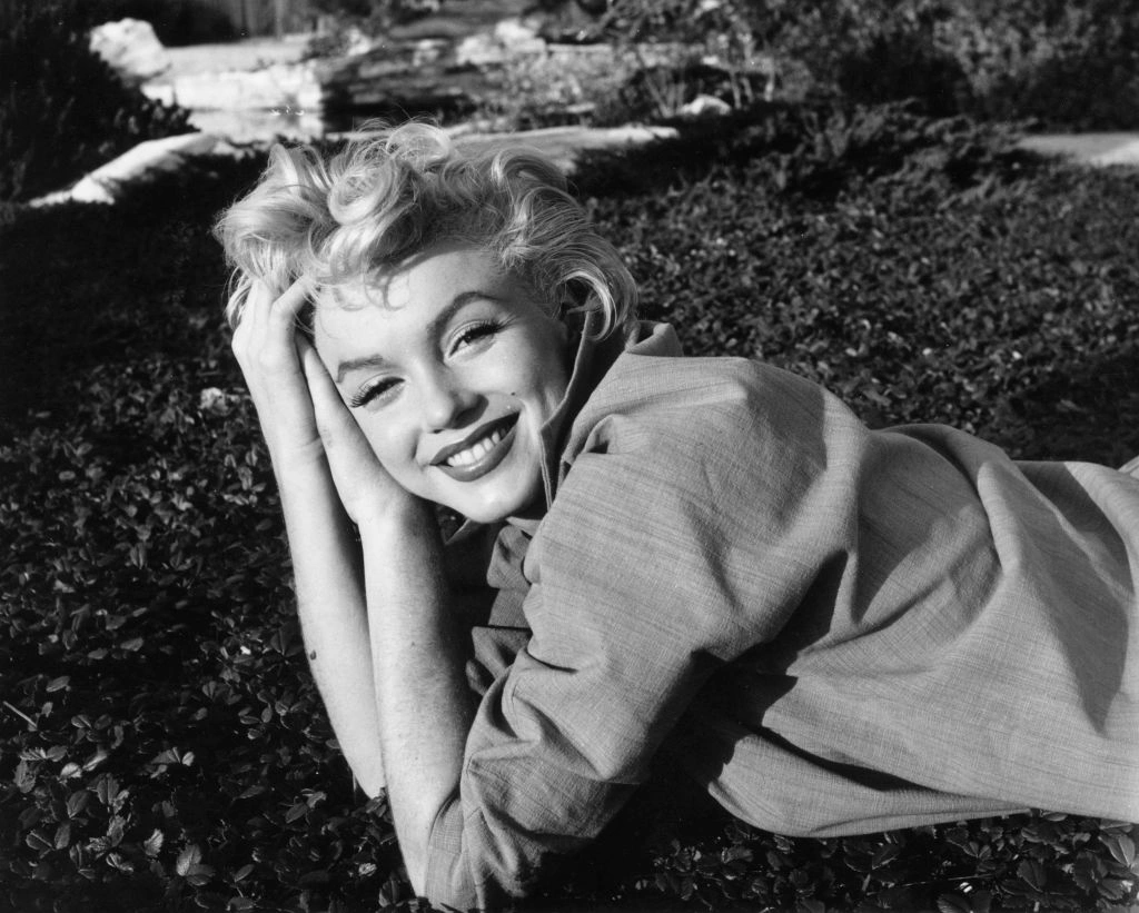 Siyah-beyaz çekilmiş, açık alanda uzanırken kameraya gülümseyen sarışın Hollywood yıldızının portresi; 1950’ler sinema estetiğini ve zamansız yıldız cazibesini yansıtan ikonik fotoğraf.