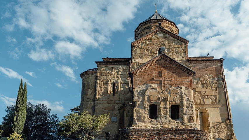 Mavi ve bulutlu bir gökyüzünün altında, Tiflis'teki tarihi Metechi Kilisesi, yıpranmış taş duvarları, tuğla işçiliğindeki haç deseni ve görkemli kubbesiyle dikkat çekerek yükselirken, solunda uzun selvileri barındıran yeşil ağaçlar görülmektedir.