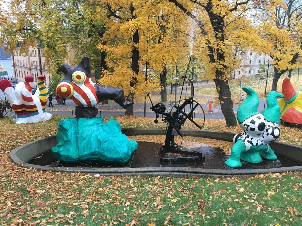 Stockholm'ün Skeppsholmen Adası'nda yer alan Moderna Museet'in bahçesinde, sonbaharın sarı yapraklarıyla kaplı zeminde, çizgili ve benekli, mizahi figürlerden oluşan modern sanat heykelleri bir su havuzunun içinde, sarı ağaçların ve tarihi bir binanın eşliğinde sergileniyor.