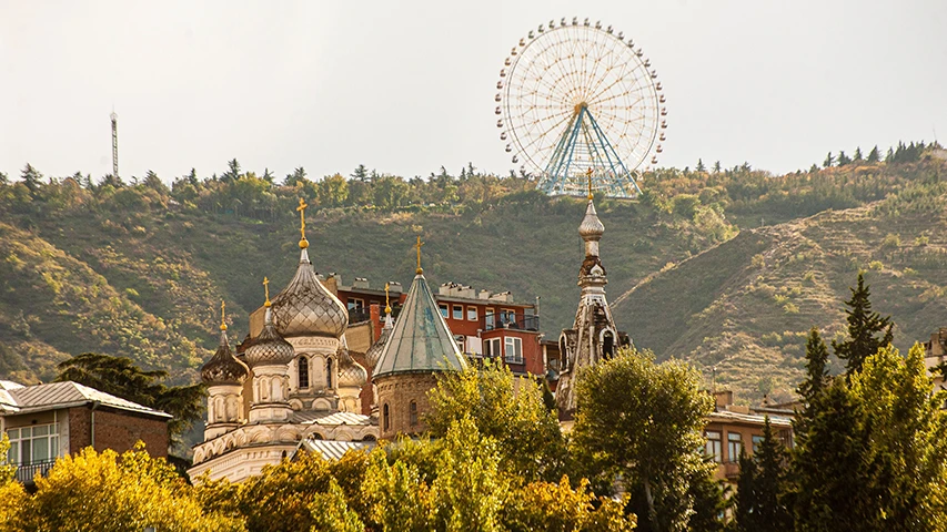 Tiflis'in yemyeşil tepelerinden birinde konumlanan Mtatsminda Park'ın ikonik dönme dolabı, ön plandaki tarihi kilise kubbeleri ve sonbahar renklerindeki ağaçlarla birlikte şehrin kültürel ve eğlence dolu yüzünü gözler önüne seriyor.