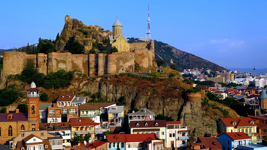 Tiflis'in sarp bir tepesi üzerine kurulu, taş duvarları ve sivri kubbeli kilisesiyle dikkat çeken görkemli Narikala Kalesi, altında uzanan rengarenk çatılı tarihi evleri, arkasında şehrin modern kuleleri ve sağda ilerleyen teleferik hattıyla panoramik bir şehir manzarasını sunuyor.