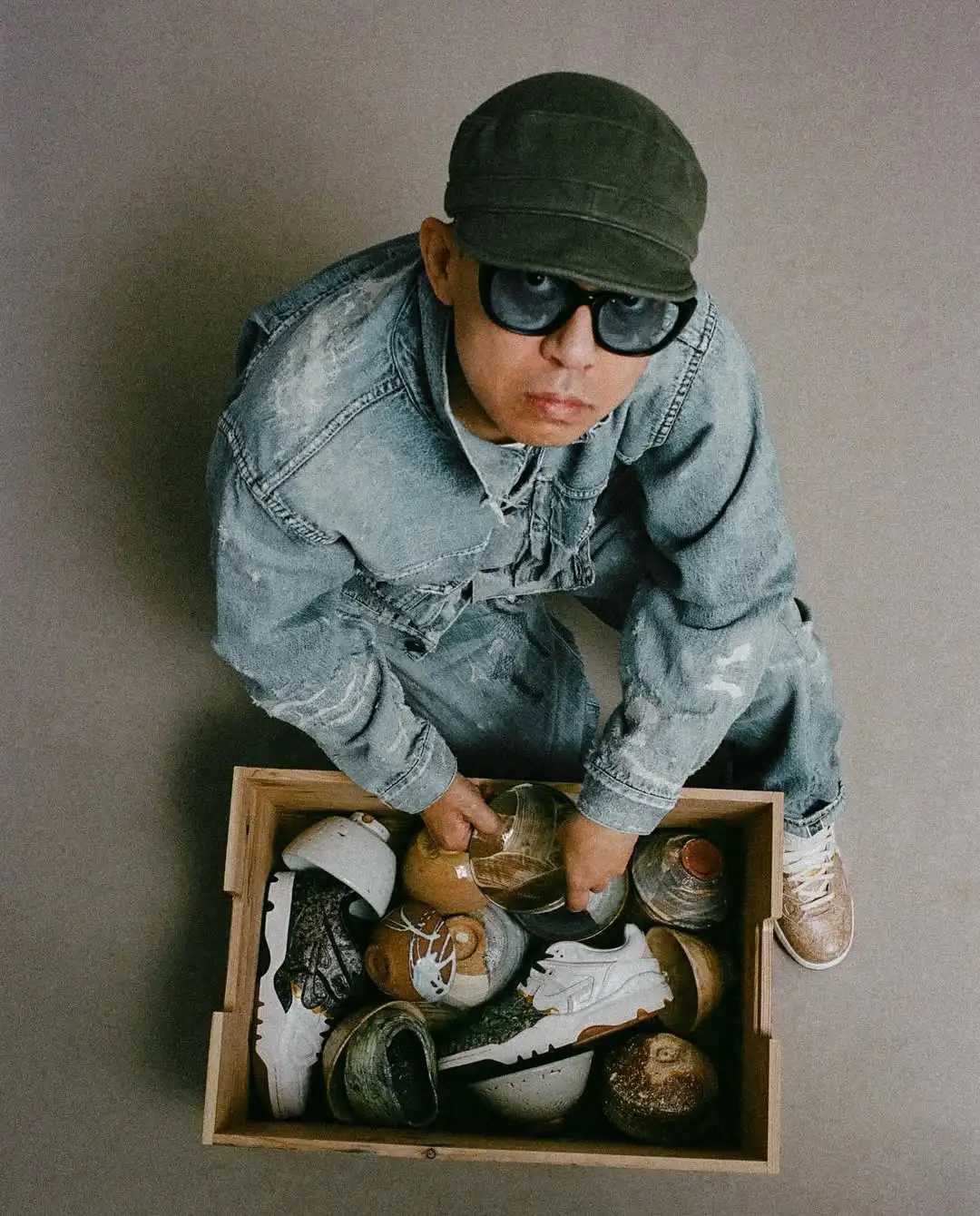 Denim ceket ve pantolonla stilize edilen, şapka ve koyu renk güneş gözlüğü takan moda tasarımcısının; el yapımı seramik objeler ve sneaker’larla dolu ahşap bir sandık başında yukarıdan çekilmiş portresi.