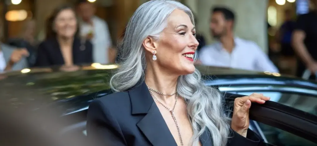 Şehirde arabadan inerken gülümseyen şık gri saçlı kadın; sürdürülebilir moda ve zamansız stil vurgusu yapan sokak stili sahnesi.