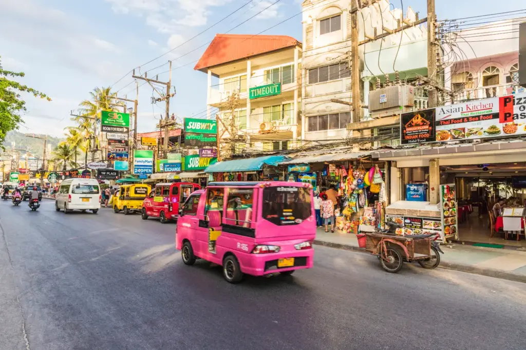 Phuket'in hareketli sokaklarında, rengarenk dükkan ve restoran tabelalarıyla çevrili bir yolda ilerleyen pembe bir tuk-tuk ve diğer araçlar, adanın dinamik ulaşım ve keşfedilecek yerler atmosferini yansıtıyor.