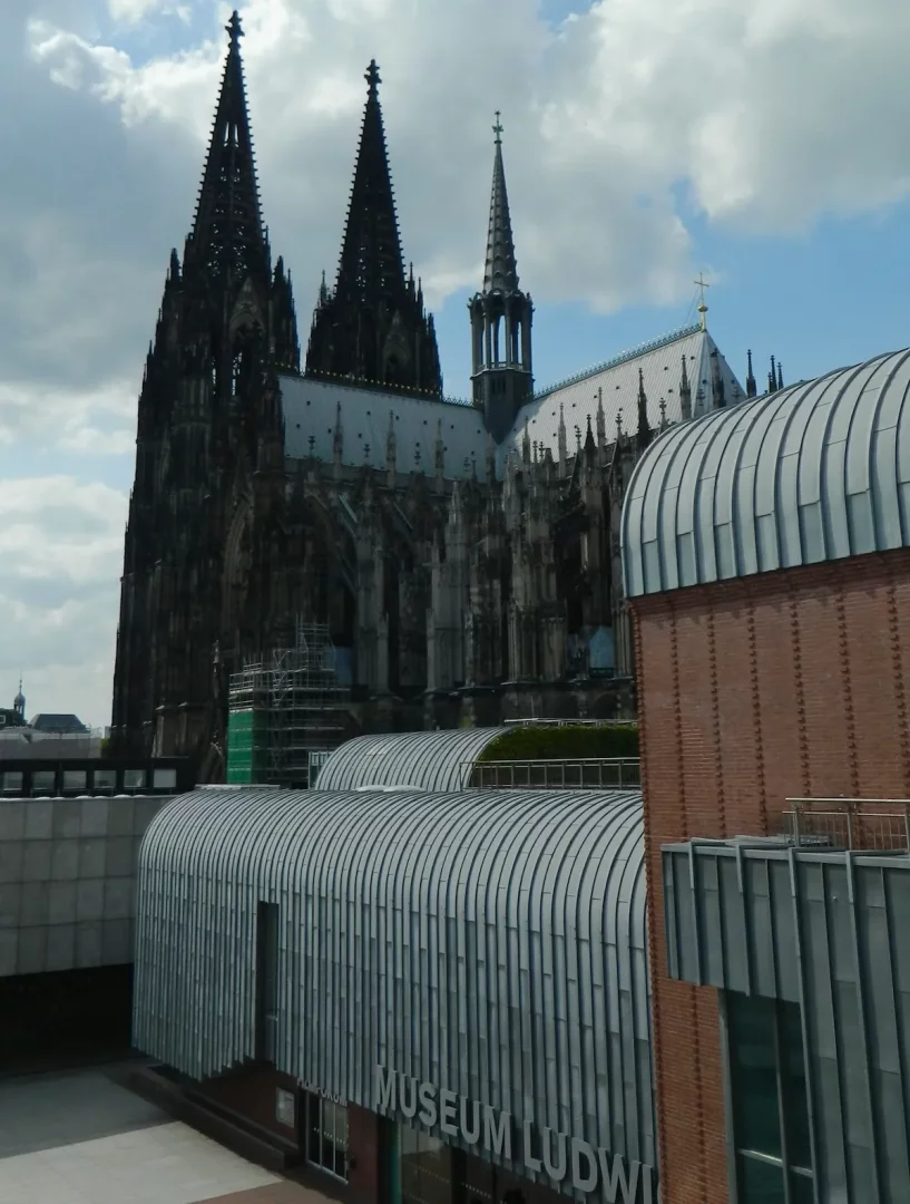 Kavisli, nervürlü metal çatısıyla modern bir görünüme sahip Museum Ludwig binası, fonunda gri gökyüzüne uzanan etkileyici Köln Katedrali'nin gotik kuleleriyle birlikte gözüküyor.