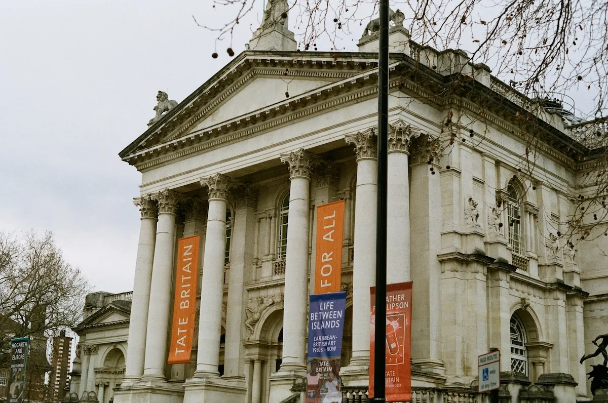 Bulutlu gökyüzünün altında, Tate Britain Müzesi'nin heybetli neoklasik cephesi, süslü sütunları ve çatıdaki heykelleriyle ön plana çıkarken, cephesinden sarkan turuncu ve mavi sergi afişleri sanat meraklılarını çeşitli eserleri keşfetmeye davet ediyor.