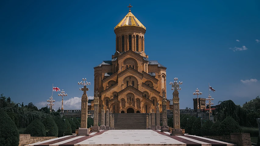Tiflis'teki Sameba Katedrali, masmavi bir gökyüzünün altında, göz alıcı altın rengi kubbesi ve detaylı mimarisiyle yükselirken, geniş basamaklı taş yolu ve zarif işlemeli sütunlarıyla çevrili bir meydanda bütün heybetiyle duruyor ve Gürcistan bayrağı uzakta nazikçe dalgalanıyor.