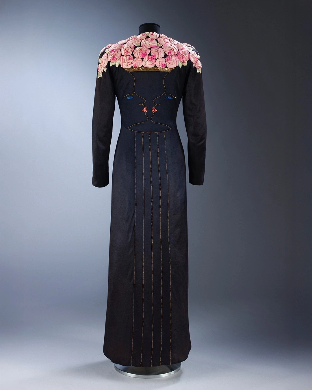 Schiaparelli’ye özgü sürrealist yüz nakışı ve omuzlarda pembe çiçek aplikeleriyle tasarlanan siyah elbise; el işçiliği detaylarıyla moda ve sanat arasındaki bağı yansıtan sergi parçası.