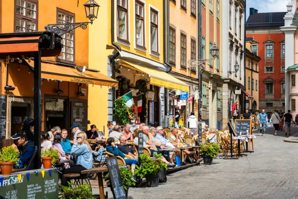 Kuzeyin En Rafine Rotası Stockholm'ün tarihi Gamla Stan bölgesinde, rengarenk Orta Çağ binalarının süslediği Arnavut kaldırımlı sokakta, yaz güneşinin altında keyifli bir Fika molası veren ve sosyalleşen çok sayıda ziyaretçi görülüyor.
