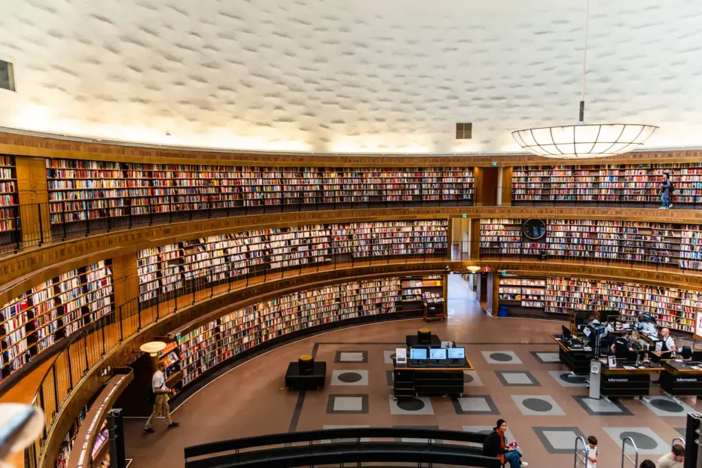 Stockholm Halk Kütüphanesi'nin ikonik silindirik ana salonu, tavanına kadar uzanan ahşap raflardaki binlerce renkli kitapla çevrili üç katmanlı dairesel yapısını gözler önüne sererken, ziyaretçiler masalarda ve orta kattaki yürüme yollarında bu etkileyici mimariyi deneyimliyor; bu eşsiz mekan, Kuzeyin en rafine rotası olan Stockholm'de gezilecek yerler arasında dikkat çekiyor.
