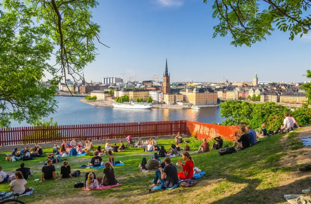 Södermalm'daki Monteliusvägen'den çekilmiş, güneşli bir yaz gününde onlarca insanın yeşil çimenlerde huzurla dinlendiği, karşıda Gamla Stan'ın tarihi yapıları, Riddarholmen Kilisesi'nin sivri kulesi ve suda süzülen bir tekneyle Stockholm'ün büyüleyici ve rafine bir gezilecek yerler manzarasını sunan keyifli bir kare.