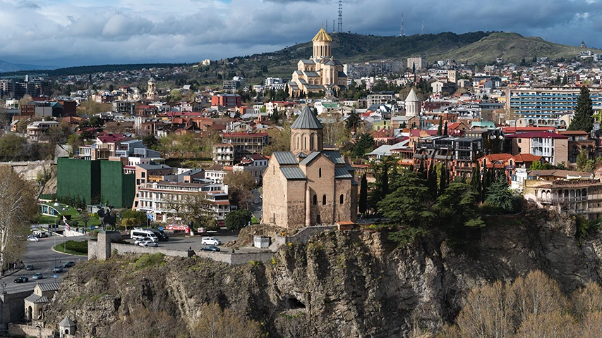 Tiflis'in etkileyici panoramik görüntüsünde, tarihi Metekhi Kilisesi kayalık bir yükseltide belirginleşirken, arka planda şehrin tepeleri boyunca uzanan yerleşim ve göz alıcı Altın Kubbeli Sameba Katedrali ile Tiflis gezilecek yerlerinin çeşitliliğini sunuyor.