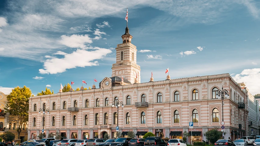 Tiflis Özgürlük Meydanı'nda, Gürcistan bayraklarıyla bezeli, pembe ve beyaz çizgili cephesi ile merkezinde saat kulesi yükselen görkemli belediye binasının önünde, parkedilmiş araçlarla dolu hareketli bir şehir meydanı manzarası.