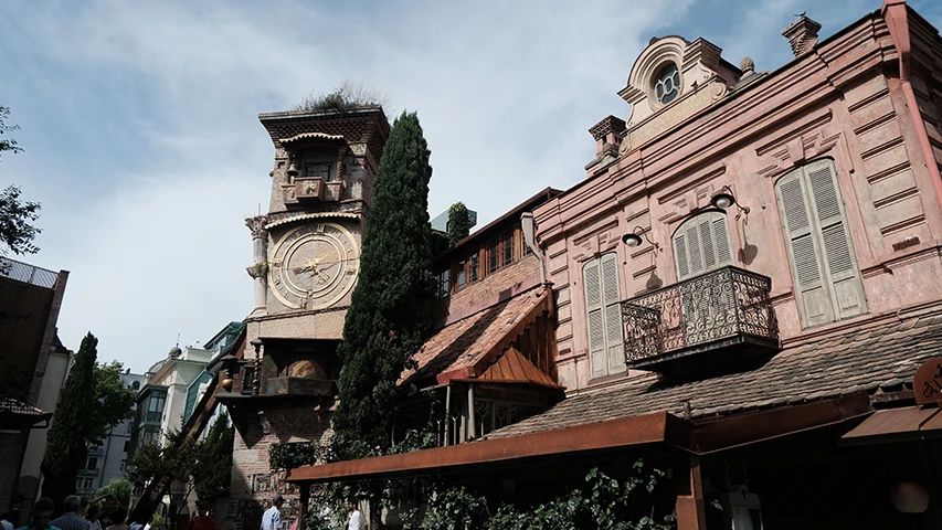 Tiflis'in tarihi atmosferini yansıtan, büyük altın rengi saat yüzüne sahip eğri duran detaylı saat kulesi, pencerelerinde panjurlar ve süslü ferforje balkonları olan pembe tonlu komşu binayla birlikte gökyüzü altında duruyor.