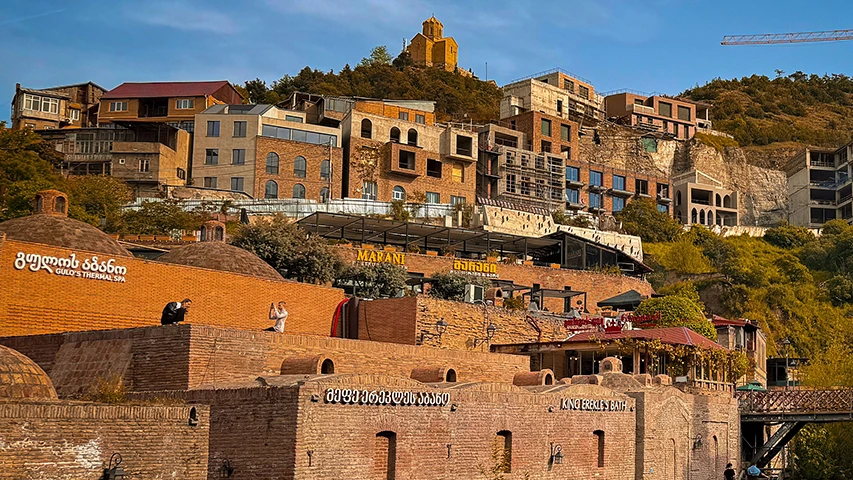 Gürcistan'ın başkenti Tiflis'in tarihi şehir merkezini gösteren bu panoramik fotoğrafta, kubbeli tuğla çatılara sahip geleneksel kükürt banyoları ön planda, yamaca doğru yükselen katmanlı yapılar ve en tepede altın rengi bir kilise gökyüzüyle buluşuyor.