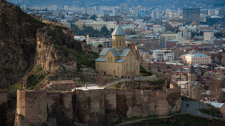 Tiflis'in tarihi güzelliklerini gözler önüne seren bu geniş açılı çekimde, yemyeşil tepelerin ve kayalıkların arasında yükselen, konik çatılı bej rengi bir kilise ile eski taş surlar, arka planda modern şehir dokusuyla birleşerek görülmeye değer bir manzara oluşturuyor.