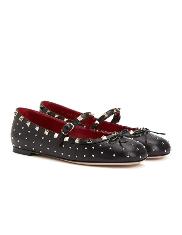 Valentino'nun Rockstud koleksiyonundan, parlak altın rengi piramit zımbalar, kırmızı iç astar, zarif bir Mary Jane kayışı ve fiyonk detayıyla süslenmiş, zamansız ve ikonik siyah deri babetler, beyaz bir zeminde şıkça sergileniyor.