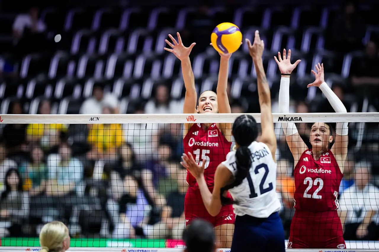 Voleybol sahasında, filenin üzerinde yükselen sarı mor topa kırmızı milli takım formasını giyen 15 numaralı oyuncu smaç basmaya çalışırken, beyaz formasıyla rakip takımın 12 numaralı oyuncusu topu bloklamak için ellerini uzatıyor ve arkada 22 numaralı diğer oyuncu da blok pozisyonunda hazır bekliyor.