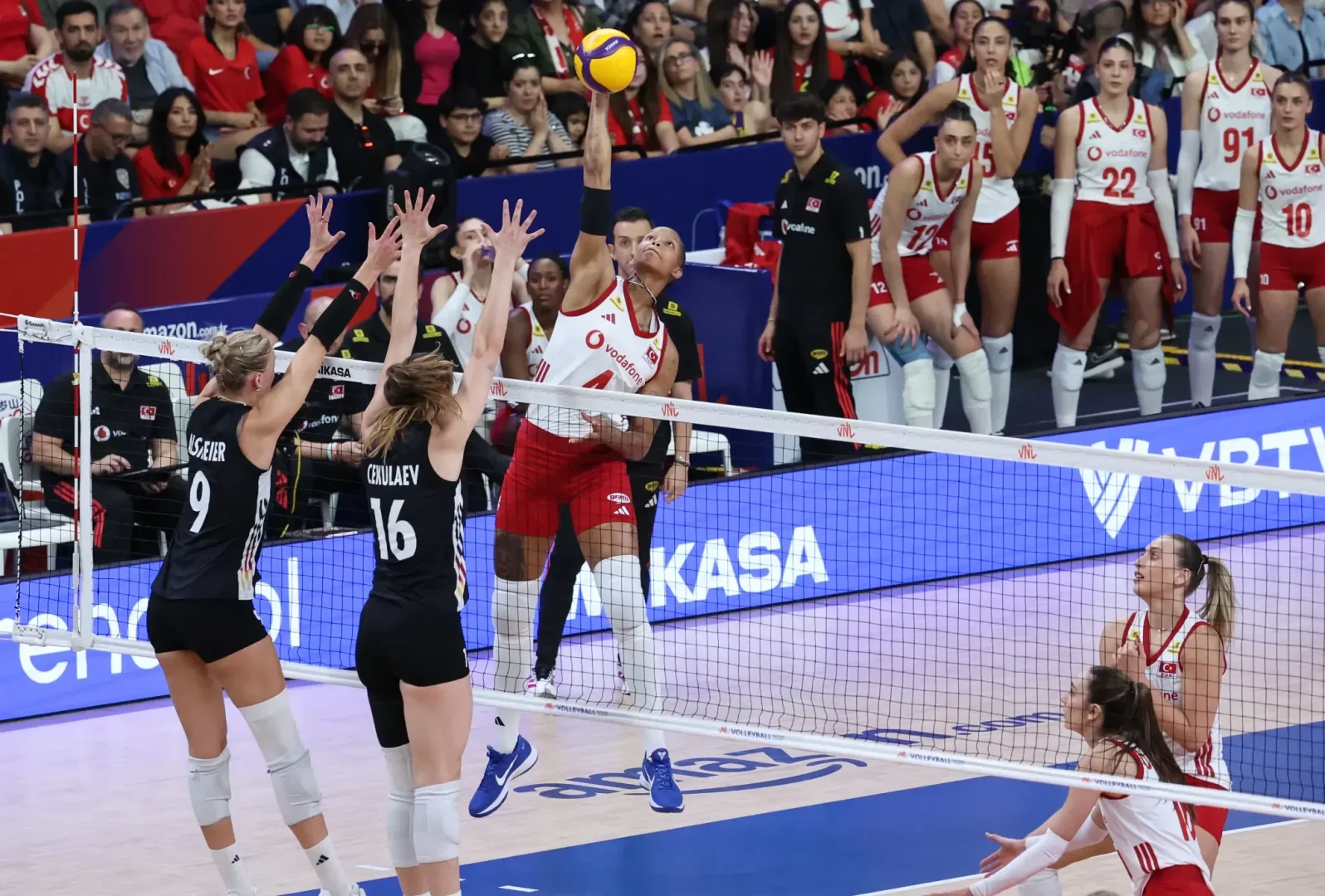 Orta sahada file üzerindeki sarı-mavi voleybol topuna güçlü bir şekilde sıçrayarak hücum vuruşu yapan kırmızı beyaz formalı kadın voleybolcu, iki siyah formalı rakip oyuncunun yükselen blok çabalarına rağmen topu karşı sahaya göndermeye çalışıyor ve bu an voleybol hücum kuralları kapsamında bir atak pozisyonunu vurguluyor.