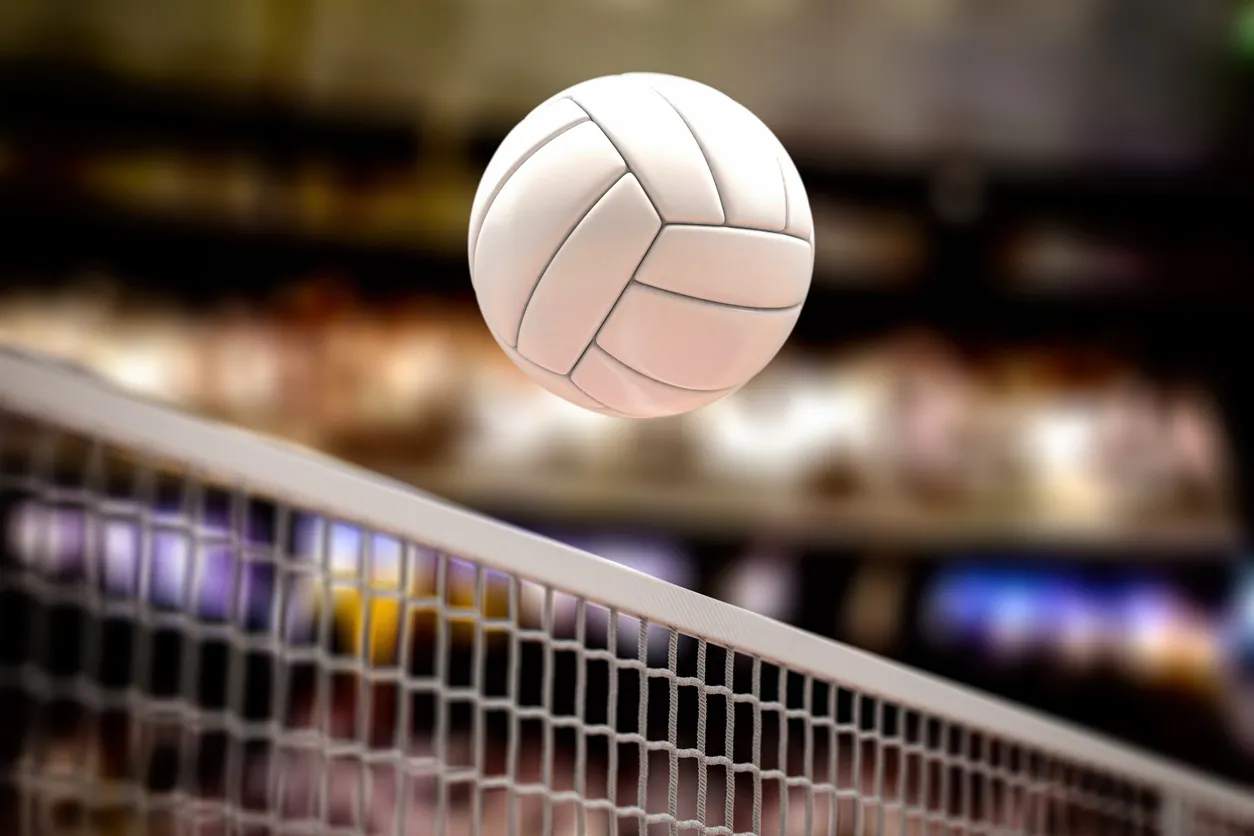 File seviyesinin üzerinde havada asılı duran beyaz bir voleybol topu, arka planda bulanık tribün ışıklarıyla bir kapalı spor salonunda voleybol kuralları ve FIVB hakkında bilgilendiren bu makaleyi destekliyor.