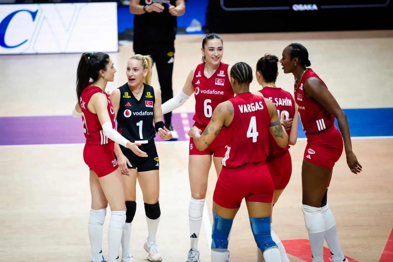 Kırmızı ve siyah formalar giyen altı Türk voleybol oyuncusu, kapalı bir kortta maç sırasında bir araya gelmiş, sarı saçlı bir oyuncu (forma 1) jestlerle konuşurken diğer takım arkadaşları dikkatle dinleyerek galibiyet için saha içi stratejilerini ve voleybol oyuncu pozisyonlarını gözden geçiriyor.
