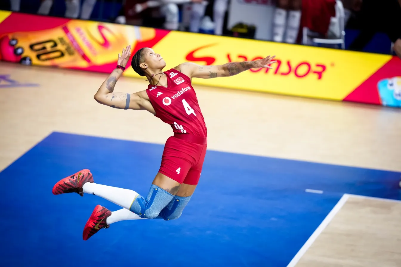 Kırmızı 4 numaralı forması, şortu ve dizlikleriyle, sağ kolu yukarıda voleybol servis atışı için gerilmiş, mavi zeminli salonda zıplayan bir kadın voleybolcu; arkasında bulanık sarı sponsor panosu seçiliyor.