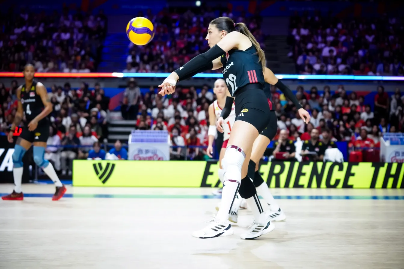 Kapalı bir voleybol maçında 19 numaralı siyah forma ve kolluklar giyen bir kadın voleybolcu, coşkulu kalabalığın önünde havada asılı duran sarı-mor topa voleybol kurallarına uygun şekilde kol pası atarak kritik bir set anına damgasını vuruyor.