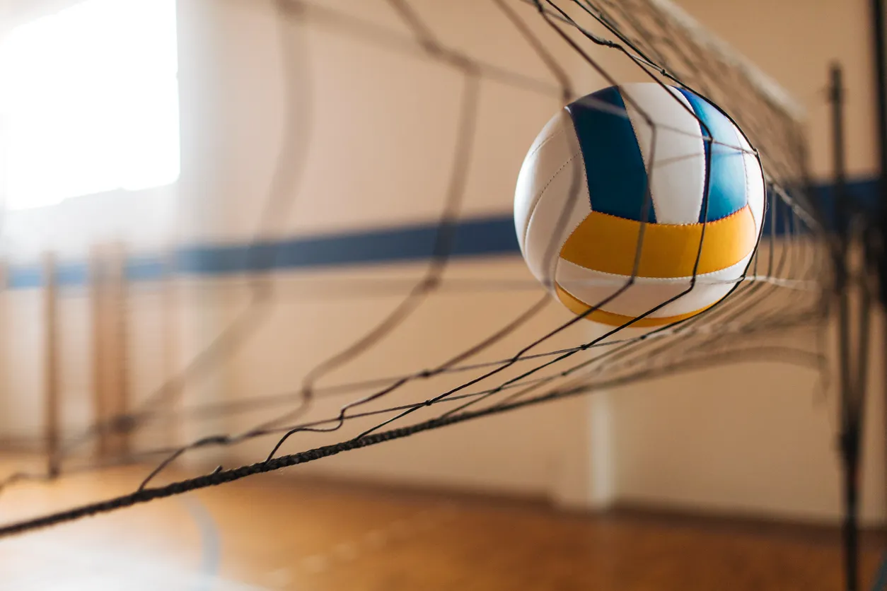 Bir spor salonunda, beyaz, mavi ve sarı renkli bir voleybol topunun fileye takılı kalarak ışıklı bir pencere önünde durduğu bu fotoğraf, voleybolda yapılan hatalar ve teknik eksiklikler hakkında düşünceler uyandırıyor.