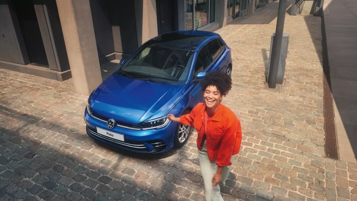 Mavi renkli Volkswagen Polo, gündüz vakti, şehir sokaklarında, önünde turuncu ceketli kadın