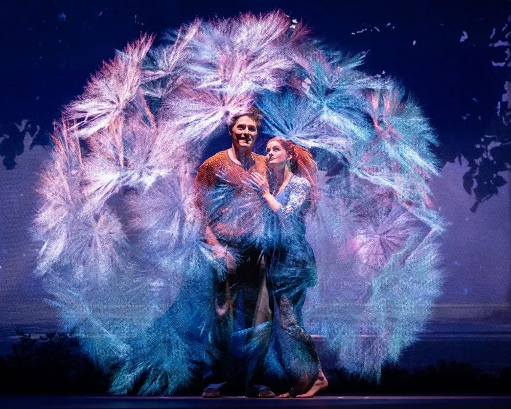 Cirque du Soleil’nin ALIZÉ gösterisinden bir sahnede, iki akrobat mavi ve mor tonlarda ışık efektleriyle çevrili bir kompozisyon içinde sahnede yan yana duruyor.