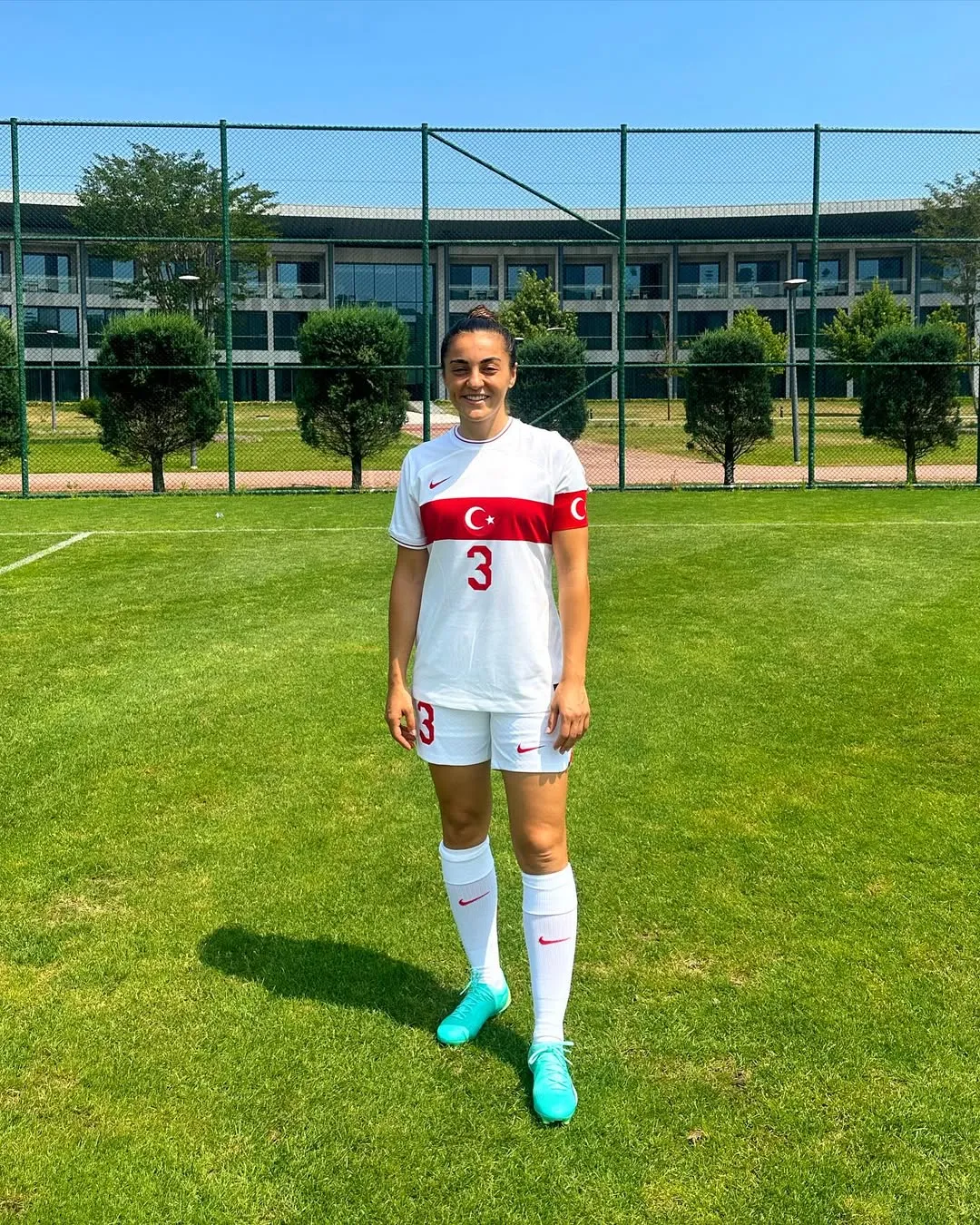 Türk milli kadın futbolcu Didem Karagenç, yeşil çim sahada beyaz milli takım forması, şortu ve turkuaz kramponlarıyla, formadaki ay yıldız ve 3 numarasıyla gülümseyerek kameraya bakıyor, arka planda tel örgü ve bir bina yer alıyor.