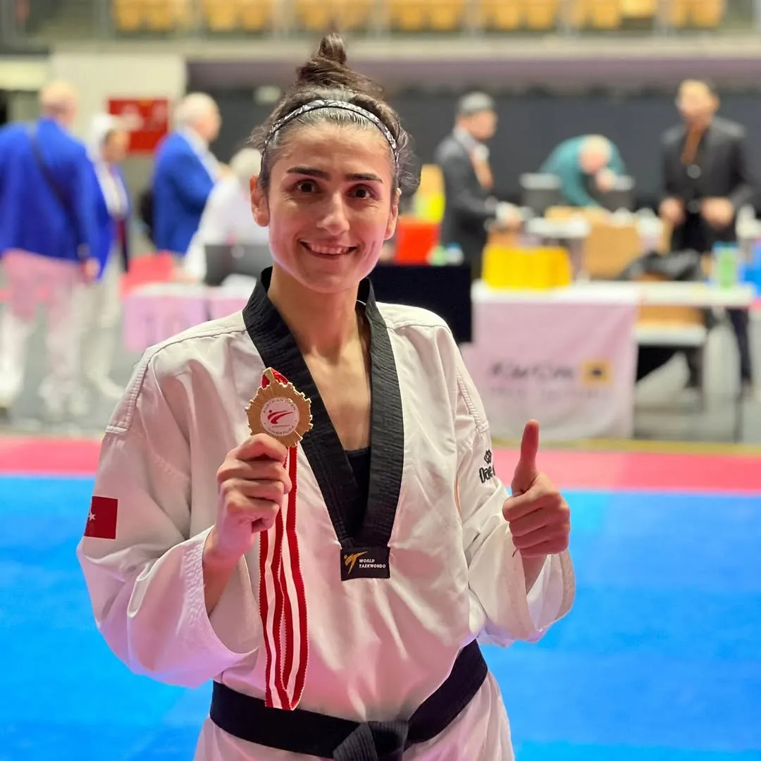 Hatice Kübra İlgün, beyaz taekwondo kıyafeti ve siyah kuşağıyla gülümsüyor, bir müsabakadan sonra gururla kazandığı altın madalyasını sol elinde tutarak sağ eliyle başparmağını yukarı kaldırıyor.