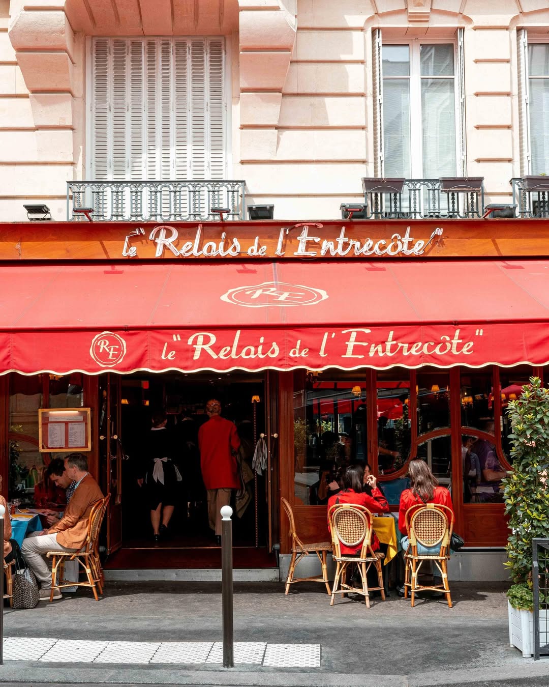 Paris’in ikonik restoranlarından Le Relais de L’Entrecôte’un Saint-Germain'deki orijinal şubesinin kırmızı tenteli, bej cepheli ve klasik brasserie atmosferini yansıtan ön görünüşü; dışarıda hasır sandalyelerde oturan müşteriler ve içeriden çıkan garsonlarla dolu, geleneksel Fransız lezzetlerini sunan bu popüler mekânın canlılığını gözler önüne seriyor.