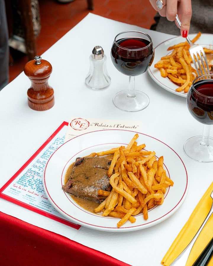 Saint-Germain'in ikonik mekanlarından Le Relais de L’Entrecôte’da beyaz masa örtüsü üzerinde, meşhur café de Paris sosuyla sunulan ince dilimlenmiş et ve altın rengi patates kızartmasıyla dolu tabaklar, iki kadeh kırmızı şarap, ahşap biberlik ve tuzluk eşliğinde, bir elin ikinci porsiyon için çatallı uzandığı iştah açıcı bir öğünü gözler önüne seriyor.