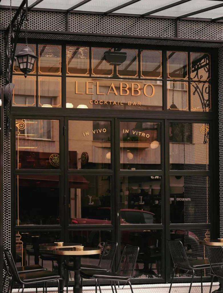Lelabbo Cocktail Bar'ın siyah çerçeveli cam cephesinde altın rengi LELABBO COCKTAIL BAR yazısı, IN VIVO ve IN VITRO ibareleri öne çıkarken, altıgen desenli kaplamaların çevrelediği girişin önünde altın tablalı yuvarlak masalar ve siyah sandalyeler zarif bir dış mekan oturma alanı sunuyor.